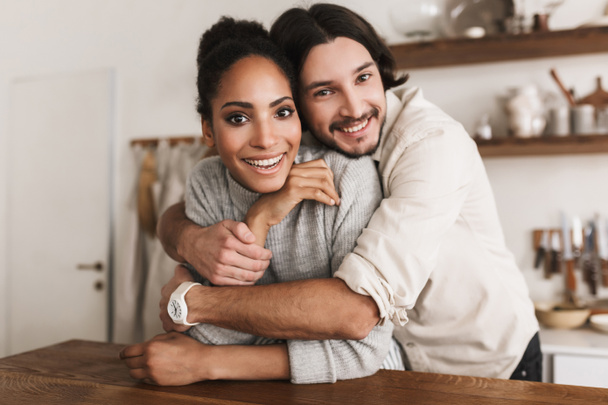 Smiling man dreamily embracing beautiful african american woman joyfully looking in camera together. Young international couple happily spending time in cozy kitchen at home - Fotó, kép