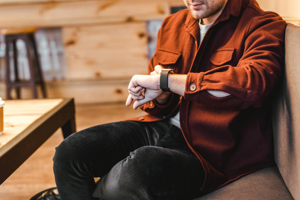 cropped view of man in burgundy shirt sitting on couch and touching watch in coffee house - Фото, изображение