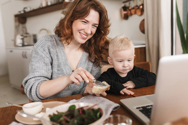 Vrolijke jongedame met rode haren in gebreide trui en zoontje zitten aan de tafel met voedsel en gelukkig kijken cartoons op laptop. Moeder tijd doorbrengen met haar baby in gezellige keuken thuis - Foto, afbeelding