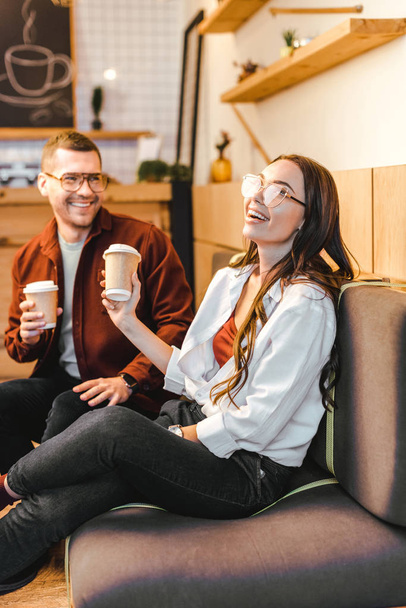 attractive woman and handsome man sitting on couch, holding paper cups and laughing in coffee house - Photo, Image