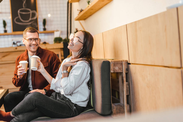 attractive woman and handsome man sitting on couch, holding paper cups and laughing in coffee house - Foto, Imagem