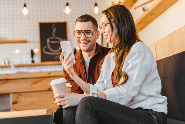 attractive woman and handsome man sitting on couch, holding paper cups and looking to smartphone in coffee house - Photo, Image
