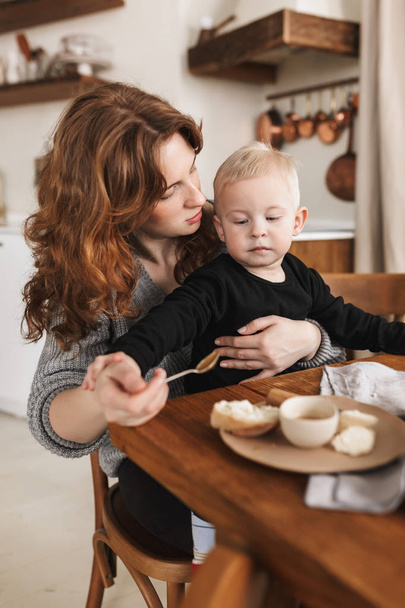 Young beautiful woman with red hair in knitted sweater sitting at the table with food dreamily feeding her little handsome son. Mom spending time with baby boy in cozy kitchen at home - Photo, Image