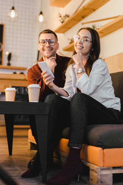 attractive woman holding smartphone and sitting on couch with handsome man at table with paper cups in coffee house - Photo, Image