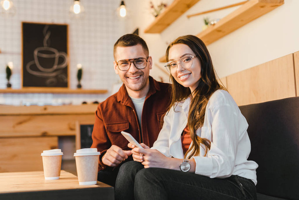 attractive woman smiling, holding smartphone and sitting on couch with handsome man at table with paper cups in coffee house - Foto, imagen