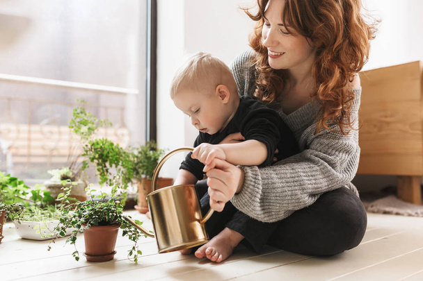 Young smiling woman sitting on floor with her little handsome son holding watering can in hands with green plants around near big window. Mom joyfully spending time with baby boy at cozy home - Photo, Image