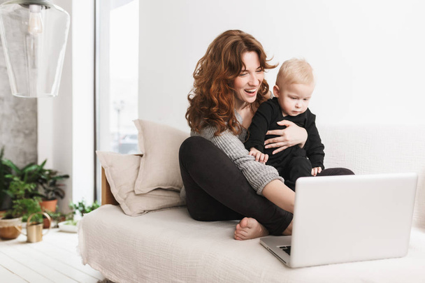 Young cheerful woman sitting on sofa with her little handsome son happily using laptop together. Mom with baby boy joyfully spending time in cozy living room at home - Photo, Image