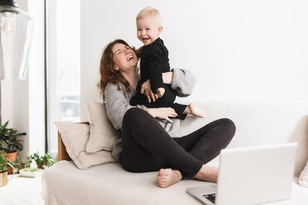 Young pretty cheerful woman sitting on sofa with her little laughing son happily playing together. Mom and baby boy joyfully spending time with laptop in cozy living room at home - Fotó, kép