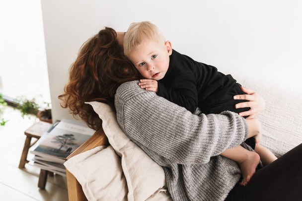 Young beautiful woman lying on sofa dreamily hugging her little handsome son that thoughtfully looking in camera. Mom with baby boy spending time in cozy living room at home - Photo, Image