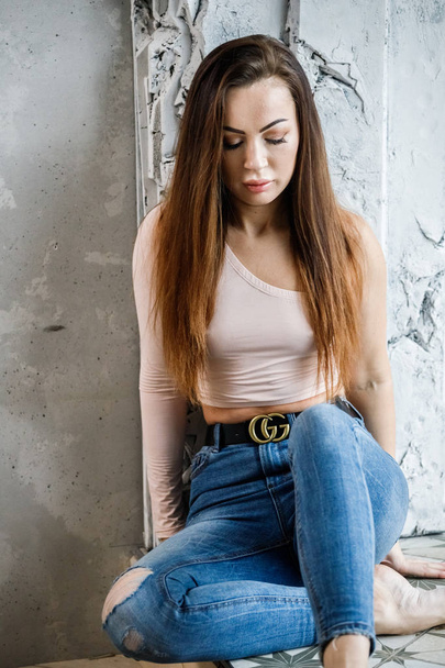 Portrait of beautiful young woman in blue jeans and beige t-shirt. Young model sitting on floor close to wall at luxury apartments interior - Foto, Imagen