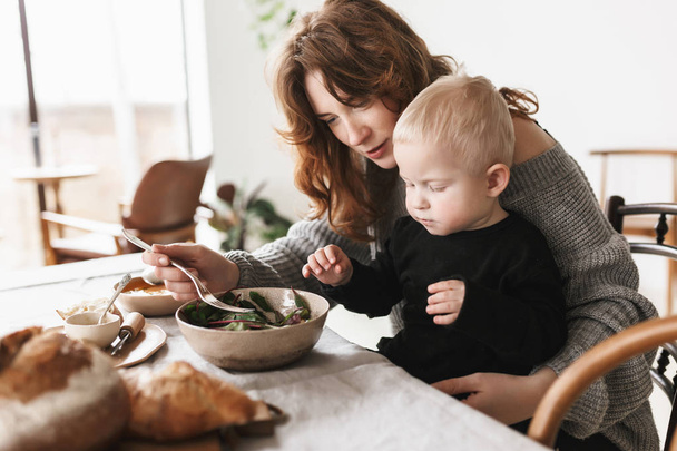 Young beautiful woman with red hair in knitted sweater sitting at the table dreamily feeding her little son. Mom spending time with baby boy in cozy kitchen at home - Photo, Image