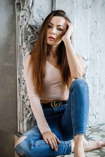 Portrait of beautiful young woman in blue jeans and beige t-shirt. Young model sitting on floor close to wall at luxury apartments interior - Fotografie, Obrázek