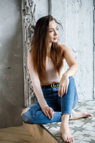 Portrait of beautiful young woman in blue jeans and beige t-shirt. Young model sitting on floor close to wall at luxury apartments interior - Photo, Image