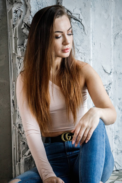 Portrait of beautiful young woman in blue jeans and beige t-shirt. Young model sitting on floor close to wall at luxury apartments interior - Fotó, kép
