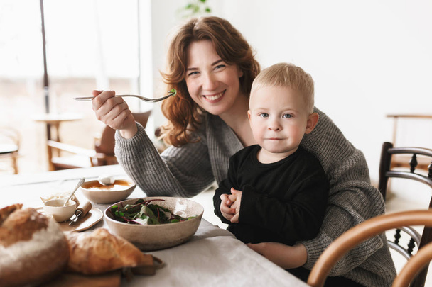 Young beautiful smiling mom with red hair in knitted sweater sitting at the table feeding her little handsome son happily looking in camera together spending time in cozy kitchen at home - Foto, Imagem