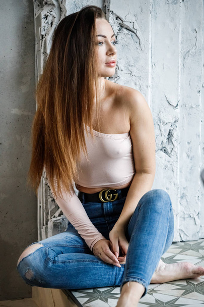 Portrait of beautiful young woman in blue jeans and beige t-shirt. Young model sitting on floor close to wall at luxury apartments interior - Foto, afbeelding