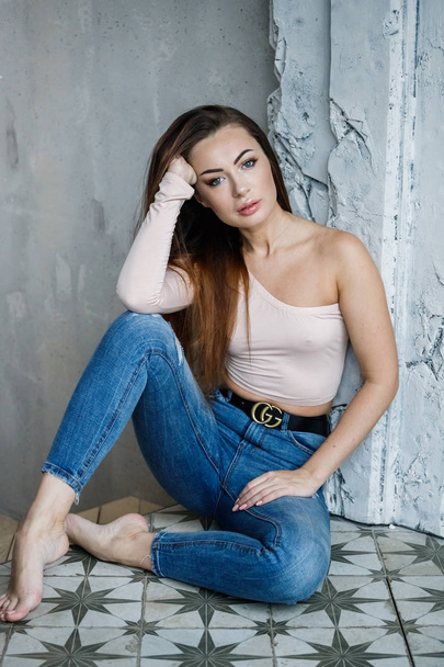 Portrait of beautiful young woman in blue jeans and beige t-shirt. Young model sitting on floor close to wall at luxury apartments interior - Photo, Image