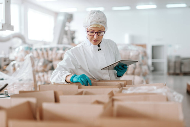 Młody pracownik Kaukaska w sterylnych uniform trzymając tablet i liczenie produktów w pudełkach. Żywność fabryka wnętrz. - Zdjęcie, obraz