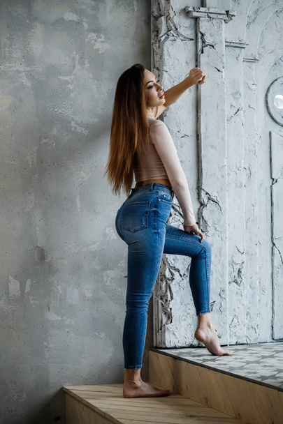 Portrét krásné mladé ženy v modré džíny a tričko béžové. Mladý model stál v blízkosti zdi v interiéru luxusní byty - Fotografie, Obrázek
