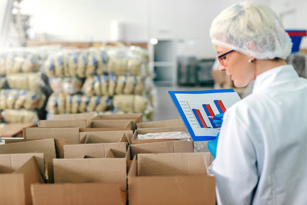 Impiegata donna in uniforme sterile e con grafico in mano contando prodotti in scatola. Interno della fabbrica alimentare
. - Foto, immagini