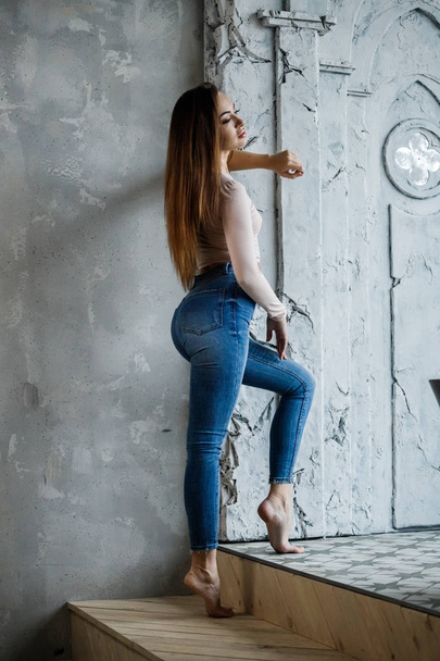 Портрет красивої молодої жінки в блакитних джинсах і бежевій футболці. Молода модель, що стоїть близько до стіни в інтер'єрі розкішних квартир
 - Фото, зображення