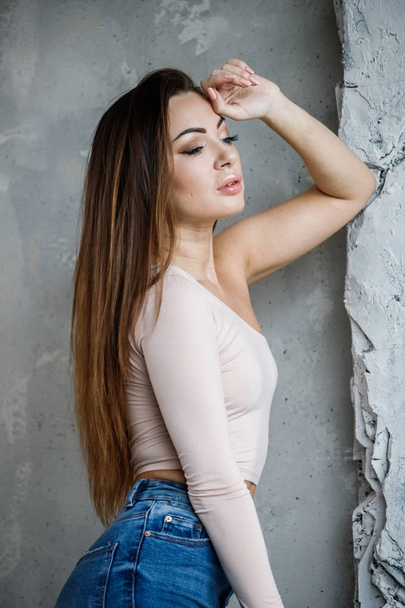 Portrait of beautiful young woman in blue jeans and beige t-shirt. Young model standing close to wall at luxury apartments interior - Φωτογραφία, εικόνα