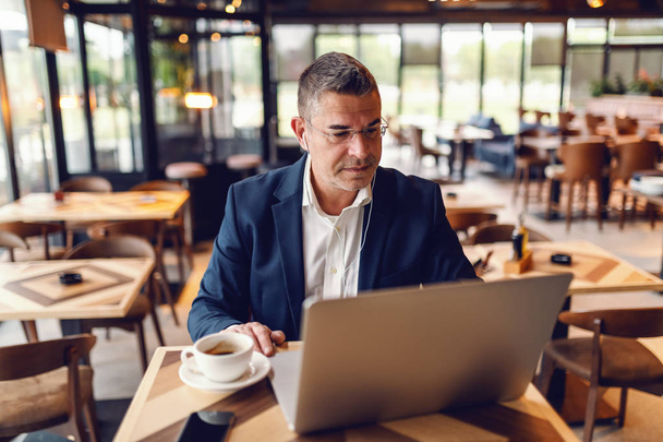 Middle aged Caucasian businessman with serious facial expression using laptop while sitting in cafe. On the desk coffee, in ears earphones. - Foto, Bild