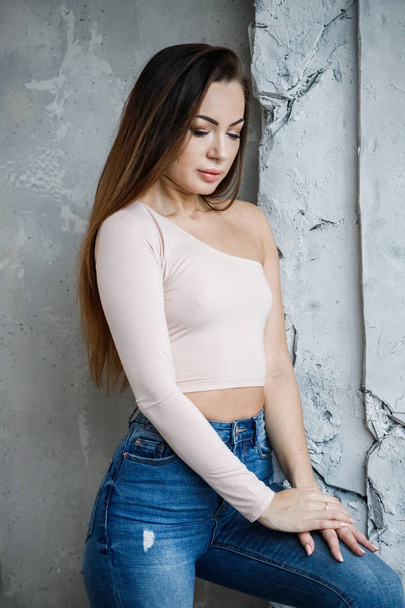 Portrait of beautiful young woman in blue jeans and beige t-shirt. Young model standing close to wall at luxury apartments interior - 写真・画像