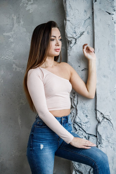Portrait of beautiful young woman in blue jeans and beige t-shirt. Young model standing close to wall at luxury apartments interior - Fotó, kép