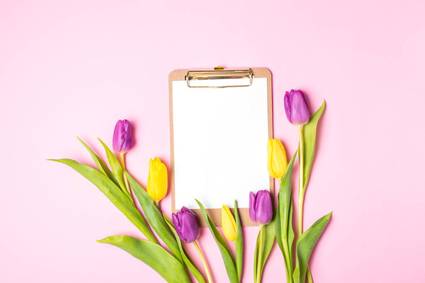 Womens Mother Day Konzept Frühlingsblumen, Lifestyleblog-Attrappe - gelbe und violette Tulpen, Klemmbrett mit leerem Papier, auf rosa Hintergrund, Kopierraum, Ansicht von oben - Foto, Bild