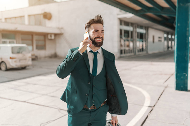 Три четверти успешного бизнесмена в формальной одежде с помощью смартфона и с багажом во время поездки на вокзал
. - Фото, изображение