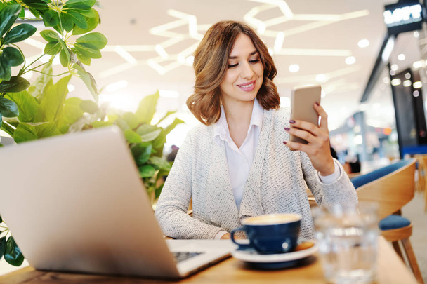 Улыбающаяся деловая женщина одевается непринужденно, используя смартфон, сидя в кафетерии. На столе ноутбук и кофе. Селективный акцент на женщине
. - Фото, изображение