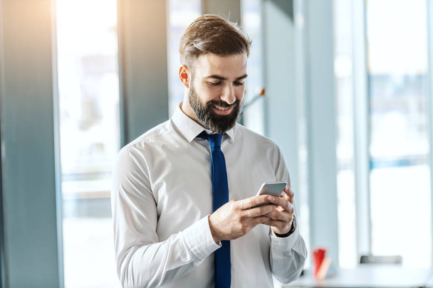 Portrait d'un beau homme d'affaires barbu et souriant en chemise et cravate en utilisant un téléphone intelligent tout en restant au bureau à côté de la fenêtre
. - Photo, image
