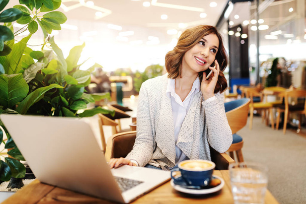 Charmante femme d'affaires souriante habillée décontractée utilisant un téléphone intelligent et un ordinateur portable tout en étant assise à la cafétéria
. - Photo, image