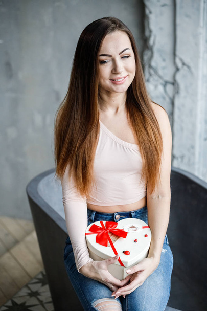 Portrait of beautiful young woman in blue jeans and beige t-shirt. Young model sitting on bath with box of sweets in hands, luxury apartments interior background  - Φωτογραφία, εικόνα