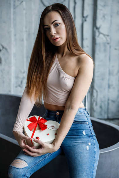 Portrait of beautiful young woman in blue jeans and beige t-shirt. Young model sitting on bath with box of sweets in hands, luxury apartments interior background  - Фото, изображение