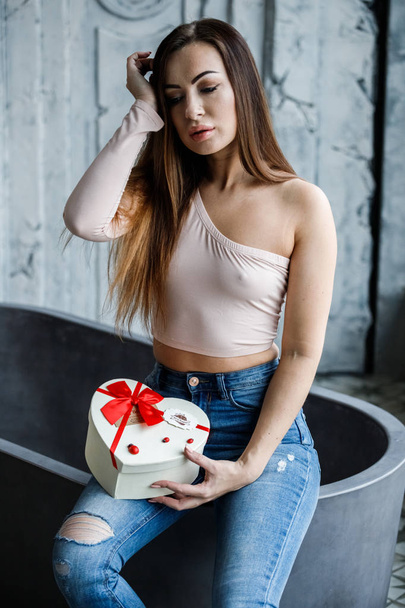 Portrait of beautiful young woman in blue jeans and beige t-shirt. Young model sitting on bath with box of sweets in hands, luxury apartments interior background  - Fotografie, Obrázek