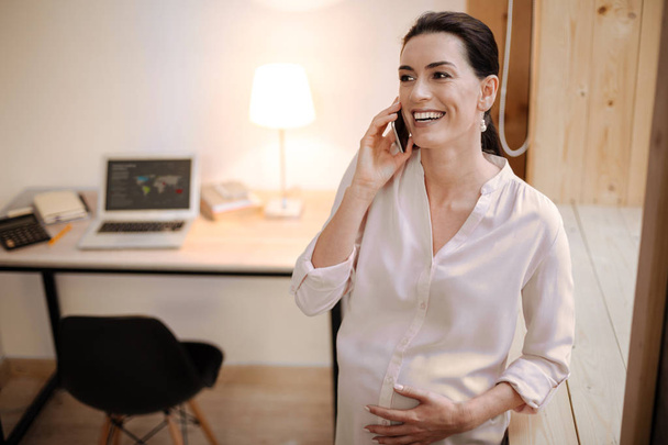 Small talk. Gay positive pregnant woman talking on phone while laughing and caressing tummy - Photo, Image