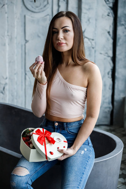 Portrait of beautiful young woman in blue jeans and beige t-shirt. Young model sitting on bath with box of sweets in hands, luxury apartments interior background  - Zdjęcie, obraz