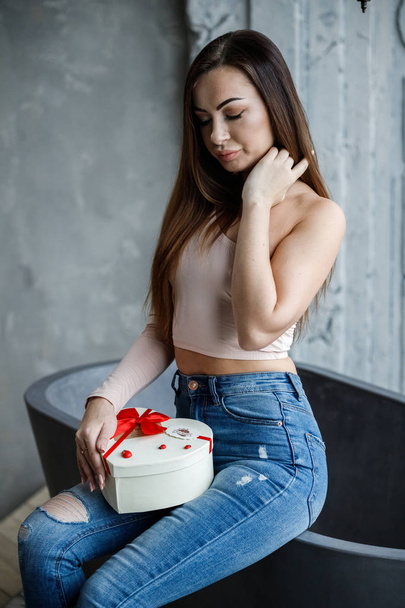 Portrait of beautiful young woman in blue jeans and beige t-shirt. Young model sitting on bath with box of sweets in hands, luxury apartments interior background  - Photo, Image