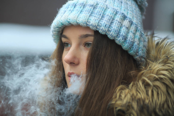Vape teenager. Young pretty white girl in blue cap is smoking an electronic cigarette on the street in the winter. Bad habit. Close up. - Foto, Imagem