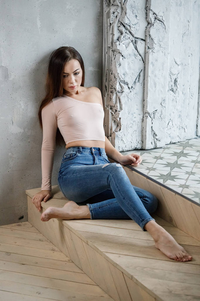 Portrait of beautiful young woman in blue jeans and beige t-shirt. Young model sitting on floor at luxury apartments interior - Photo, Image