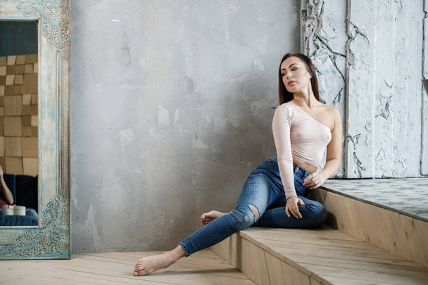 Portrait of beautiful young woman in blue jeans and beige t-shirt. Young model sitting on floor at luxury apartments interior - Fotó, kép