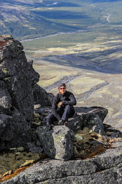 Caucasian white male tourist in sportswear, buff and glasses sits on a rock against the backdrop of mountains - Foto, Bild