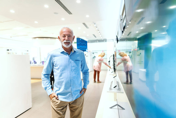 Smiling bearded senior man standing with hands in pockets. Tech store interior. - 写真・画像