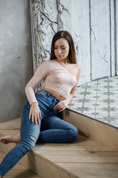 Portrait of beautiful young woman in blue jeans and beige t-shirt. Young model sitting on floor at luxury apartments interior - Foto, afbeelding