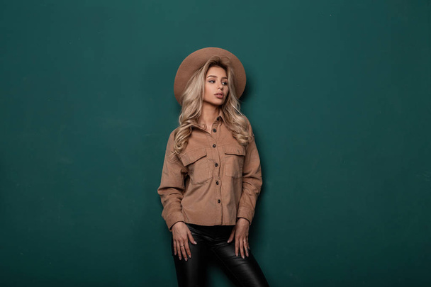 Modern fashionable woman with long blond hair in a beige elegant hat in a vintage shirt in stylish leather pants in retro style posing in a studio near a green wall. Glamor girl. - Photo, image