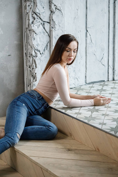 Portrait of beautiful young woman in blue jeans and beige t-shirt. Young model sitting on floor at luxury apartments interior - Фото, зображення