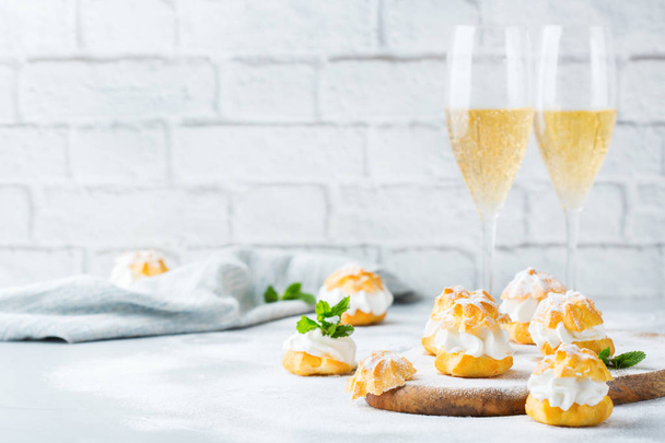 Еда и напитки, праздничная концепция. Вкусные сладкие домашние профитроли со сливками и бокалами шампанского на современном кухонном столе
 - Фото, изображение