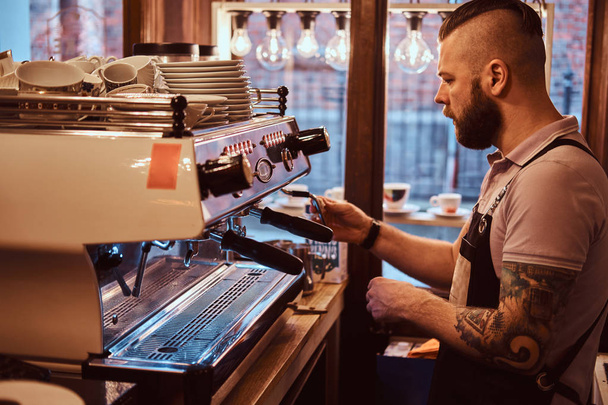 tätowierter Barista mit stylischem Bart und Frisur bei der Arbeit an einer Kaffeemaschine in einem Café oder Restaurant - Foto, Bild